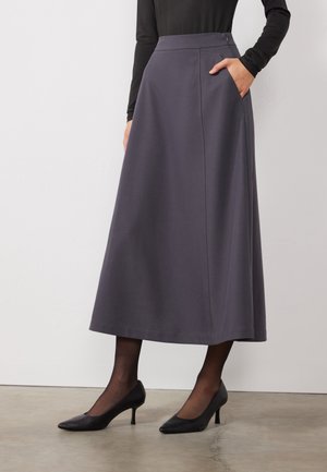 Kobieta ubrana w ciemnoszarą spódnicę midi o kroju A z wysokim stanem i bocznymi kieszeniami, w połączeniu z czarnymi rajstopami, czarnymi szpilkami oraz czarną bluzką z długim rękawem.