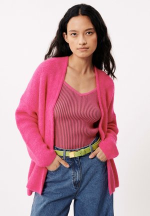 Cardigan rose flou, haut rose côtelé, ceinture verte avec boucle en métal, jeans bleu foncé amples. Textures douces et couleurs audacieuses.