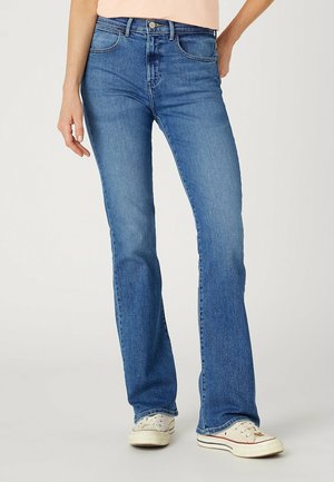 Nainen, joka käyttää puolikorkeavyötäröisiä sinisiä bootcut-farkkuja ja valkoisia tennareita, seisoo käsi lanteilla.