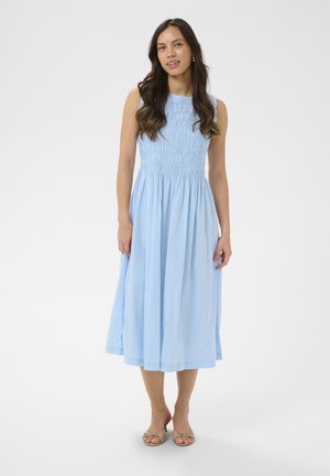 Vrouw met lang donker haar, gekleed in een lichtblauwe mouwloze midijurk en beige sandalen, staande tegen een effen witte achtergrond.