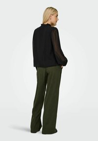 Schwarze Bluse mit Polka-Dots, transparenten langen Ärmeln und gerafftem Ausschnitt, kombiniert mit olivgrünen, weit geschnittenen Cordhosen.
