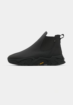 Botín negro sin cordones con un acabado liso y suela texturizada, con una pestaña para facilitar el uso. Marca Vibram visible en la suela.