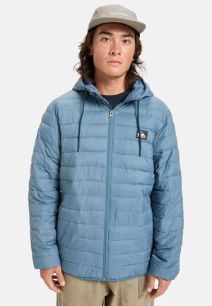 Blauwe pufferjack met horizontale gewatteerde textuur, voorrits, verstelbare capuchon en trekkoord. Draagt over een donker T-shirt.