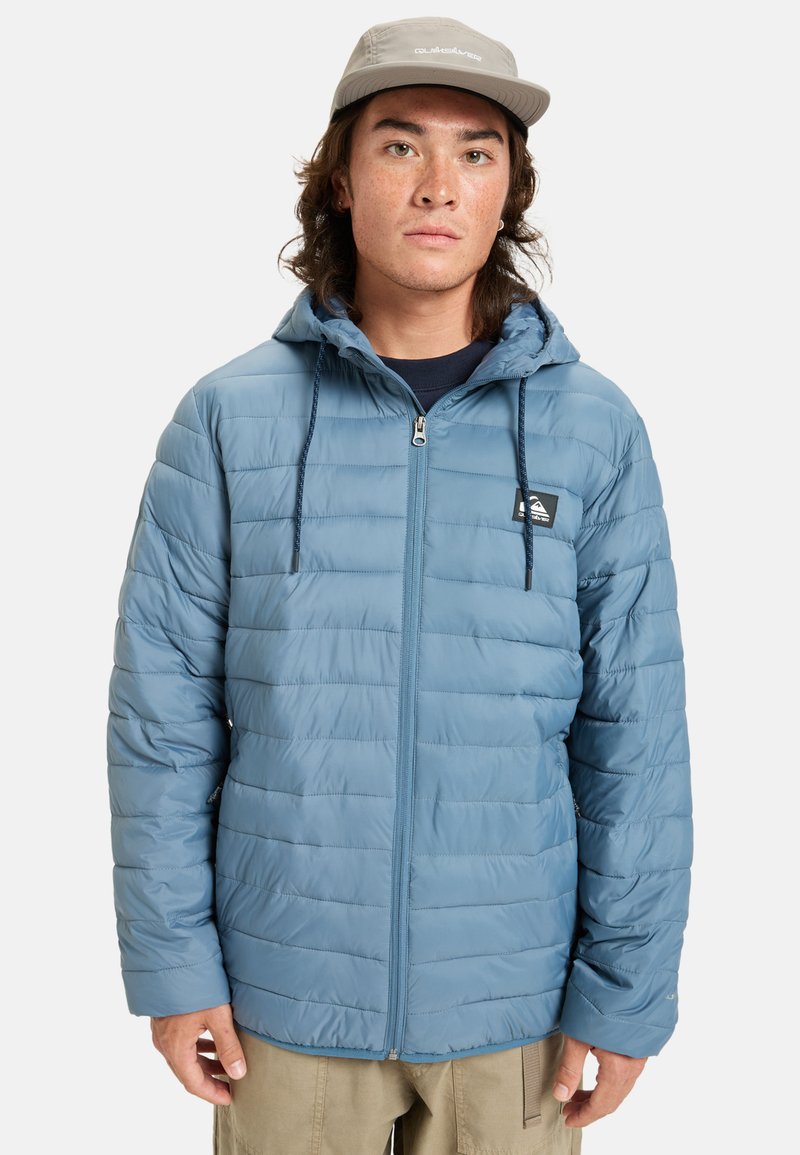 Blå puffjacka med horisontell quiltad struktur, framdragkedja, justerbar huva och dragsko. Bärs över en mörk t-shirt.