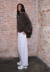 Giacca in velluto a coste marrone, pantaloni larghi da jogging grigi e sneakers argento. Capelli intrecciati sciolti, con sfondo di un muro di mattoni rustico.