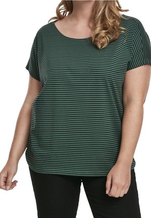 Kvinde iført en mørkegrøn og sort kortærmet t-shirt med vandrette striber og sorte bukser, stående med armene afslappet langs siderne.