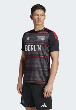 Črno-siva nogometna majica z rdečimi črtami, ki ima trikotno vzorčenje, napis 'BERLIN' in logotip. Prileganje črne kratke hlače z logotipom.