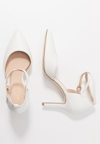 Chaussures à talons hauts blanches avec un bout pointu, une finition texturée et des brides de cheville. Elles ont une semelle intérieure beige et un talon mince.