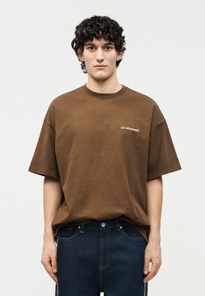 Νεαρός άνδρας με σγουρά σκούρα μαλλιά που φοράει ένα φαρδύ καφέ μπλουζάκι με το κείμενο "LES BENJAMINS" και σκούρα μπλε τζιν, στέκεται μπροστά σε ένα απλό φόντο.