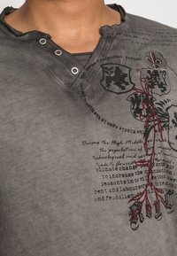 Camisa gris de manga corta con un cuello desgastado, acentos de botones y un diseño floral bordado en rojo con texto negro y elementos heráldicos.