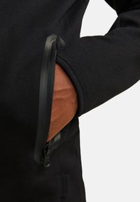 Détail d'une veste noire montrant une poche zippée avec un revêtement intérieur texturé marron. La poche possède une fermeture éclair noire et un tissu lisse.