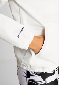 Suéter blanco con puños elásticos y un bolsillo lateral. La manga tiene un logotipo negro de "STORM". Tejido texturizado con un acabado suave.