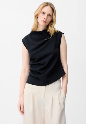 Vrouw draagt een mouwloze zwarte gedrapeerde top en lichtbeige plissébroek met hoge taille, staande met één hand in de zak tegen een witte achtergrond.