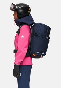 Marinblå ryggsäck med flera fack, orange detaljer och grå remmar; bärs av en person i en rosa vattentät jacka och handskar.
