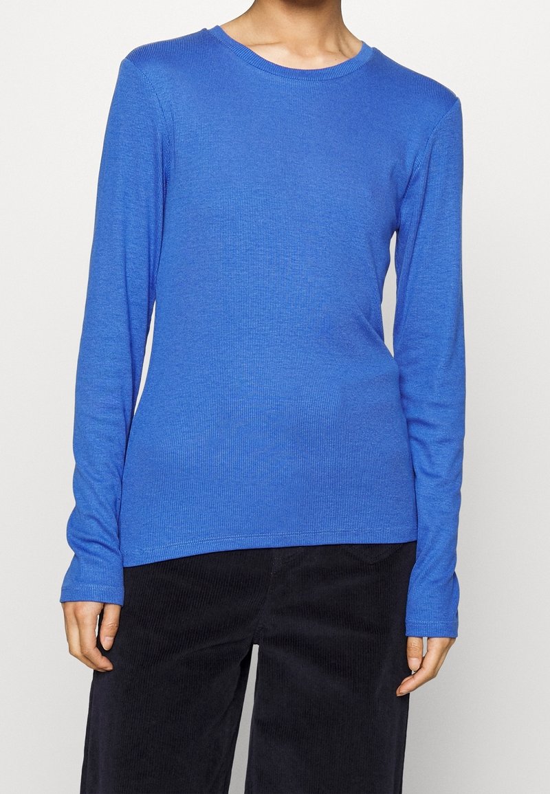 Modré tričko s dlouhým rukávem a kulatým výstřihem, žebrovaná textura, přiléhavý střih, doplněné černými manšestrovými kalhotami.