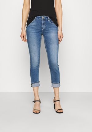 Slim-fit blauwe spijkerbroek met opgerolde pijpen, gedragen met een zwart mouwloos topje en zwarte sandalen met hoge hakken, op een lichte achtergrond.