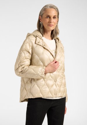 Reife Frau mit grauen Haaren, die Perlenohrringe trägt, eine hellbeige gesteppte Jacke mit Kapuze, weißes Hemd und schwarze Hose, posiert vor weißem Hintergrund.