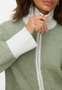 Zielona kurtka z zamkiem błyskawicznym w deseń, z białym ściągaczem przy kołnierzu i mankietach. Posiada teksturowany materiał oraz błyszczące srebrne wykończenie zamka.