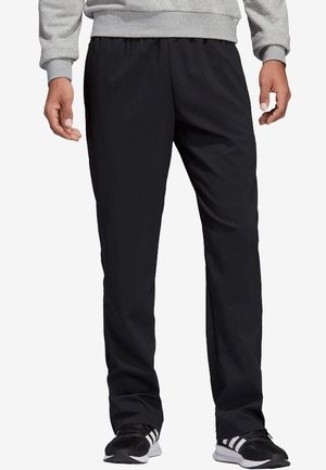 Homme portant un pantalon noir droit, un sweat gris et des chaussures de sport noires avec des bandes blanches, debout, les bras détendus.