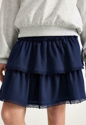 Laivastonsininen kerroksittainen hame, joka on valmistettu teksturoidusta kankaasta ja jossa on pitsireunus. Hameessa on röyhelöidyt reunat ja elastinen vyötärönauha, ja se on yhdistetty harmaaseen collegepaitaan.