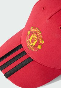 Červená baseballová čiapka s ohnutým krempou, na prednej strane so žltým logom Manchester United a tromi čiernymi pruhmi. Bavlnený materiál.