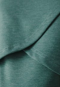 Tkanina w kolorze turkusowym o miękkiej teksturze, z nakładającymi się warstwami i widocznymi detalami szycia, wskazującymi na swobodny design odzieżowy.