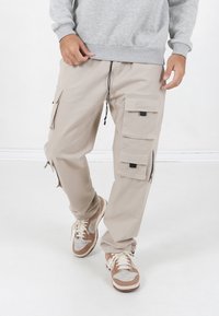 Beige cargobroek gemaakt van lichtgewicht materiaal, met meerdere zakken, een verstelbare trekkoord taille en ritsen bij de enkel.