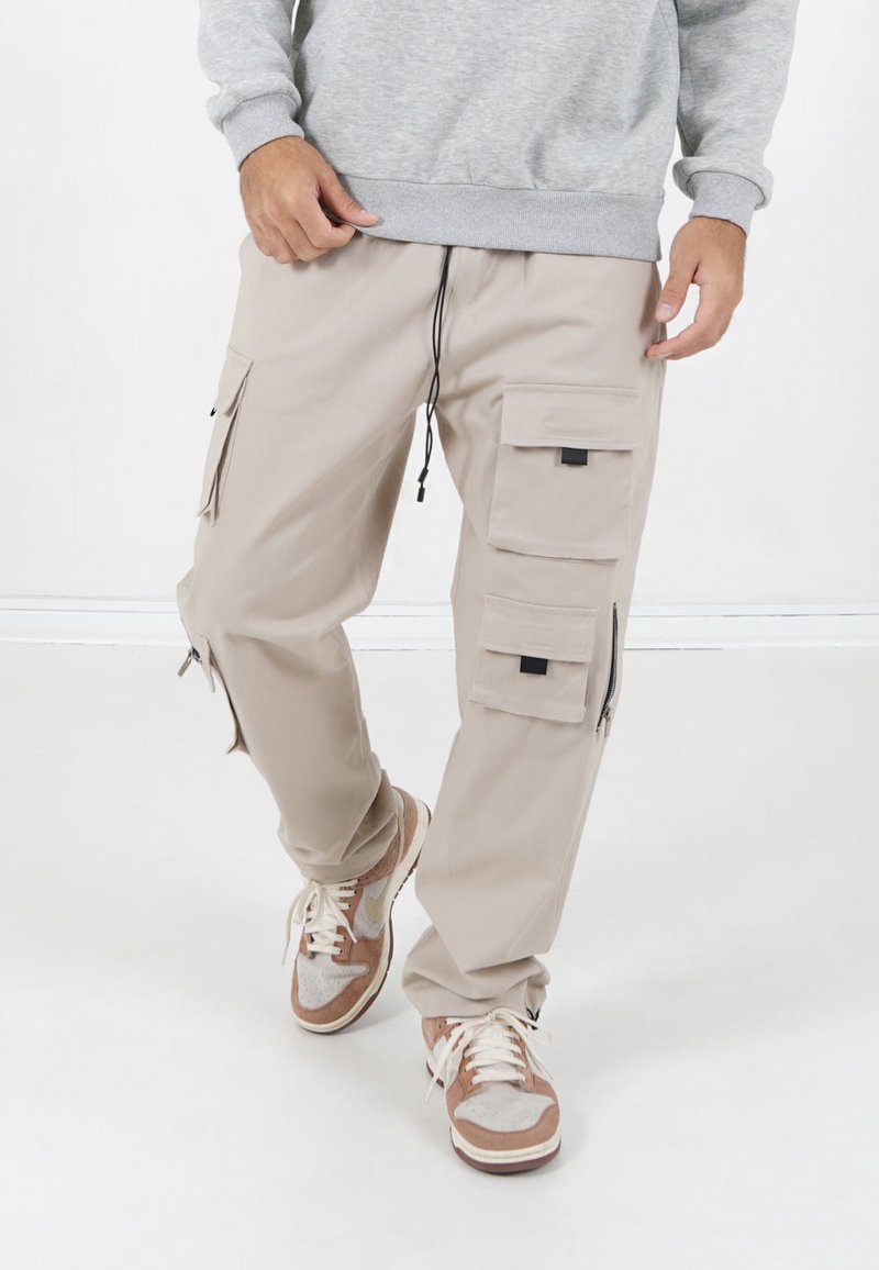 Beige cargobroek gemaakt van lichtgewicht materiaal, met meerdere zakken, een verstelbare trekkoord taille en ritsen bij de enkel.