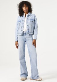 Chaqueta de denim azul claro con dos bolsillos delanteros, cierres de botón y un ajuste relajado. Combinada con unos jeans de denim azul claro de pierna ancha y deportivas beige.