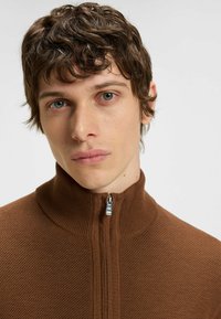 Maglione zip-up marrone con trama a rilievo e colletto alto. Presenta una cerniera metallica e dettagli a coste su tutta la superficie. Design aderente.