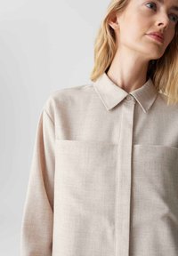 Camicia beige chiaro con colletto, chiusura a bottoni e due tasche sul petto. Il tessuto testurizzato presenta un motivo sottile.