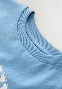 Vaaleansininen puuvillapaita, jossa on ribattu pyöreä kaula-aukko ja sileä pinta. Kankaassa on hienovarainen kuvio ja ompeludetails.