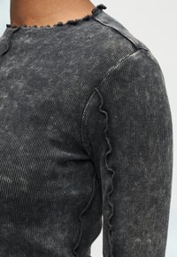 Γκρι ριμπ μακρυμάνικη μπλούζα με στρογγυλή λαιμόκοψη που έχει διακοσμητική βολάν στην άκρη. Το ύφασμα έχει απαλή, υφή με διακριτικό σχέδιο.