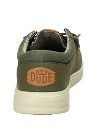 Grüner synthetischer Slipper mit einem strukturierten, bräunlichen Lederakzent, ausgestattet mit einem "HEY DUDE"-Logo auf der Rückseite und einer weißen Sohle.