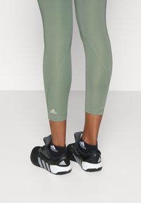 Scarpe da ginnastica nere con strisce bianche, abbinate a leggings aderenti verdi oliva; dettagli del logo evidenti su entrambi gli oggetti.