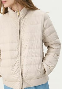 Giacca trapuntata beige con colletto alto, trapuntatura orizzontale, chiusura con zip e tasche laterali; texture del tessuto morbida e leggera.