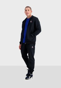 Sudadera negra con cremallera y bolsillos frontales, combinada con una camiseta azul y pantalones de chándal negros, cuenta con detalles de logo. Llevada con zapatillas deportivas en color azul marino.