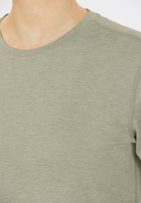 Camiseta de color oliva claro, hecha de una tela suave, que cuenta con un cuello redondo clásico y un sutil detalle de costura. Acabado texturizado con un logo en relieve.