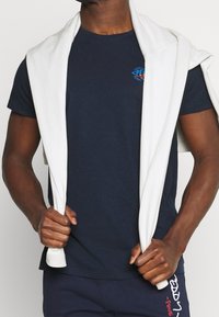 Mörkblå bomulls t-shirt med rund hals och en liten blå och röd logotyp på bröstet, tillsammans med en vit tröja draperad över axlarna.