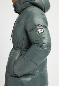 Gevoerde groene pufferjack met een hoge kraag, elastische manchetten, zijzakken en een merkembleem op de mouw. Glanzende, gladde textuur.