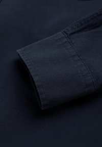 Camisa de algodón azul marino de manga larga con puños doblados, que presenta una textura suave y costuras detalladas a lo largo del dobladillo y el borde de los puños.