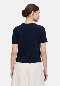 T-shirt bleu marine à manches courtes avec un col rond, texture lisse et un petit détail dans le haut du dos. L'ourlet arrive à la taille.