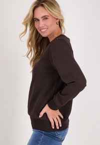 Bruine sweatshirt met lange mouwen, een ronde halslijn en geribbelde manchetten. Gepaard met lichtblauwe jeans, zijwaarts weergegeven tegen een neutrale achtergrond.