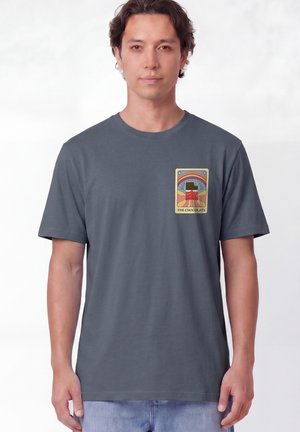 Homme portant un t-shirt gris foncé avec un petit motif coloré représentant du chocolat, un dessert et un arc-en-ciel sur le côté gauche de la poitrine, debout devant un fond blanc.