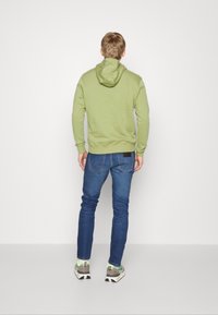 Sudadera verde con capucha ajustable, de corte relajado, combinada con vaqueros azules y zapatillas de color claro. Costuras visibles y puños acanalados.