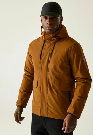 Mężczyzna ubrany w przypaloną pomarańczową kurtkę z kapturem i dużymi przednimi kieszeniami oraz czapkę bejsbolową, stojący na tle jasnego, jednolitego tła.