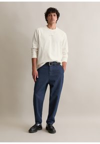 Helle beige Langarmbluse mit dezentem Text, dunkelblaue lässige Jeanshose und schwarze klobige Lederschuhe, vor einem neutralen Hintergrund.