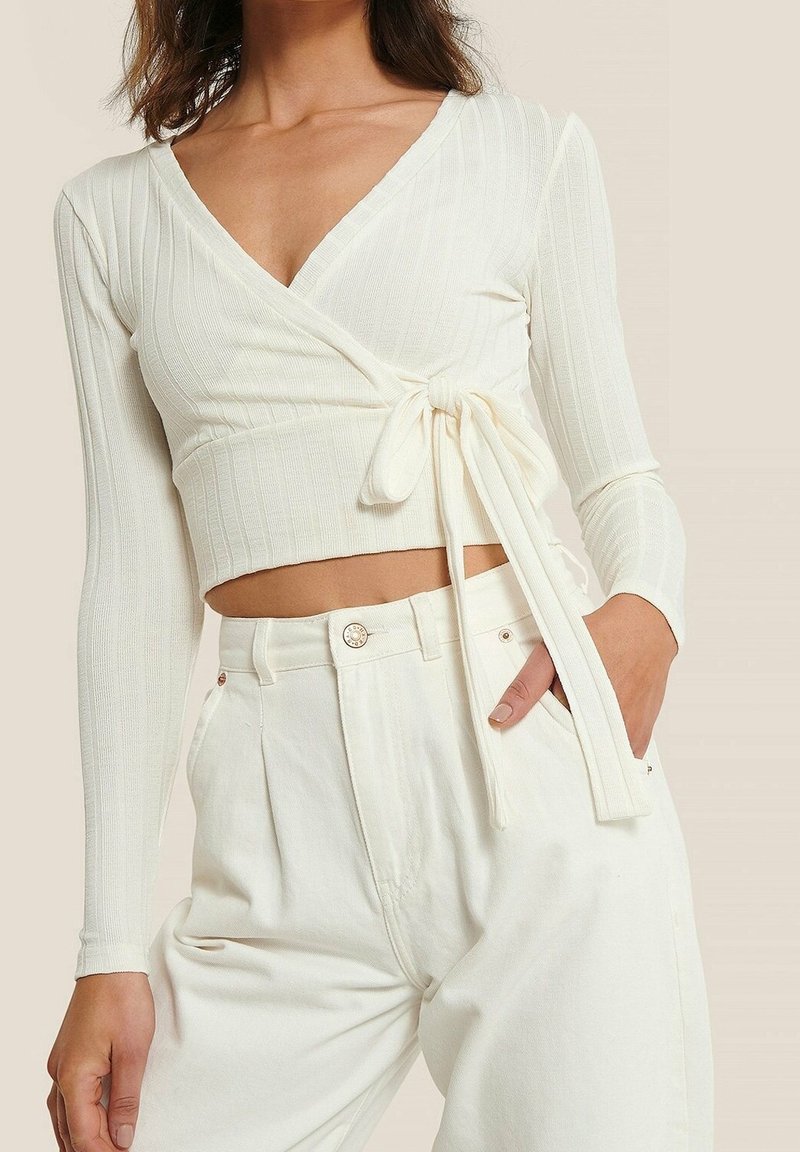 Femme portant un haut cache-cœur blanc, court, à manches longues et côtelé, noué à la taille, associé à un pantalon blanc ample à taille haute.
