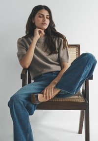 Kobieta z długimi ciemnymi włosami siedzi skrzyżowanymi nogami na drewnianym krześle z plecionym siedziskiem, ubrana w szary sweter z krótkim rękawem, niebieskie dżinsy i ciemne buty.