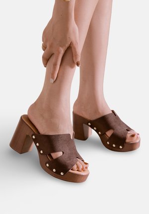 Personne portant des sandales à talons blocs ouverts en daim marron, tenant leur cheville avec des ongles de pieds et des ongles peints sur un fond uni.